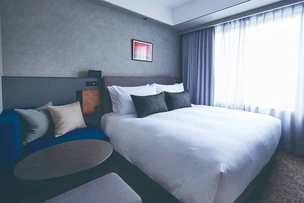Une chambre d’hôtel moderne dotée d’un grand lit, d’un coin salon avec une table ronde et d’une grande fenêtre avec des rideaux.