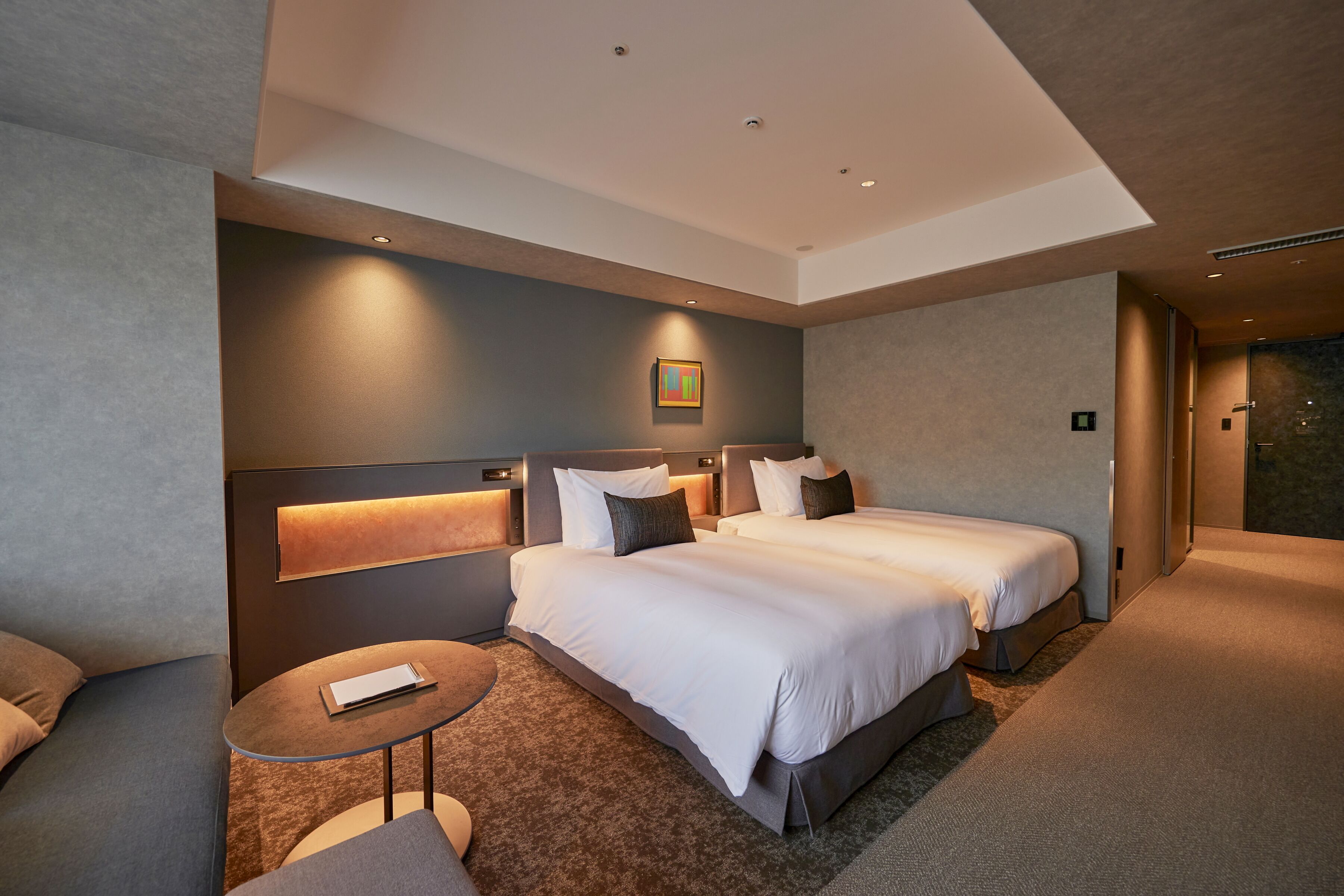Une chambre d’hôtel moderne avec deux lits, une petite table ronde et une tête de lit dotée d’un éclairage intégré.