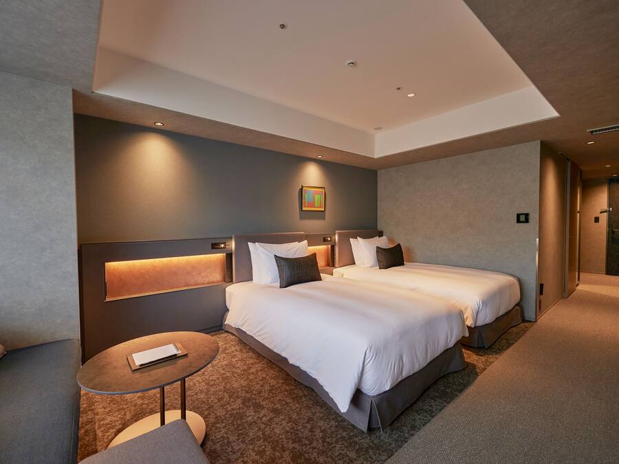 A modern hotel room with two beds, a small round table, and a headboard with built-in lighting.