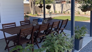 Patio esterno con tavolo e sedie di legno, circondato da piante in vaso.