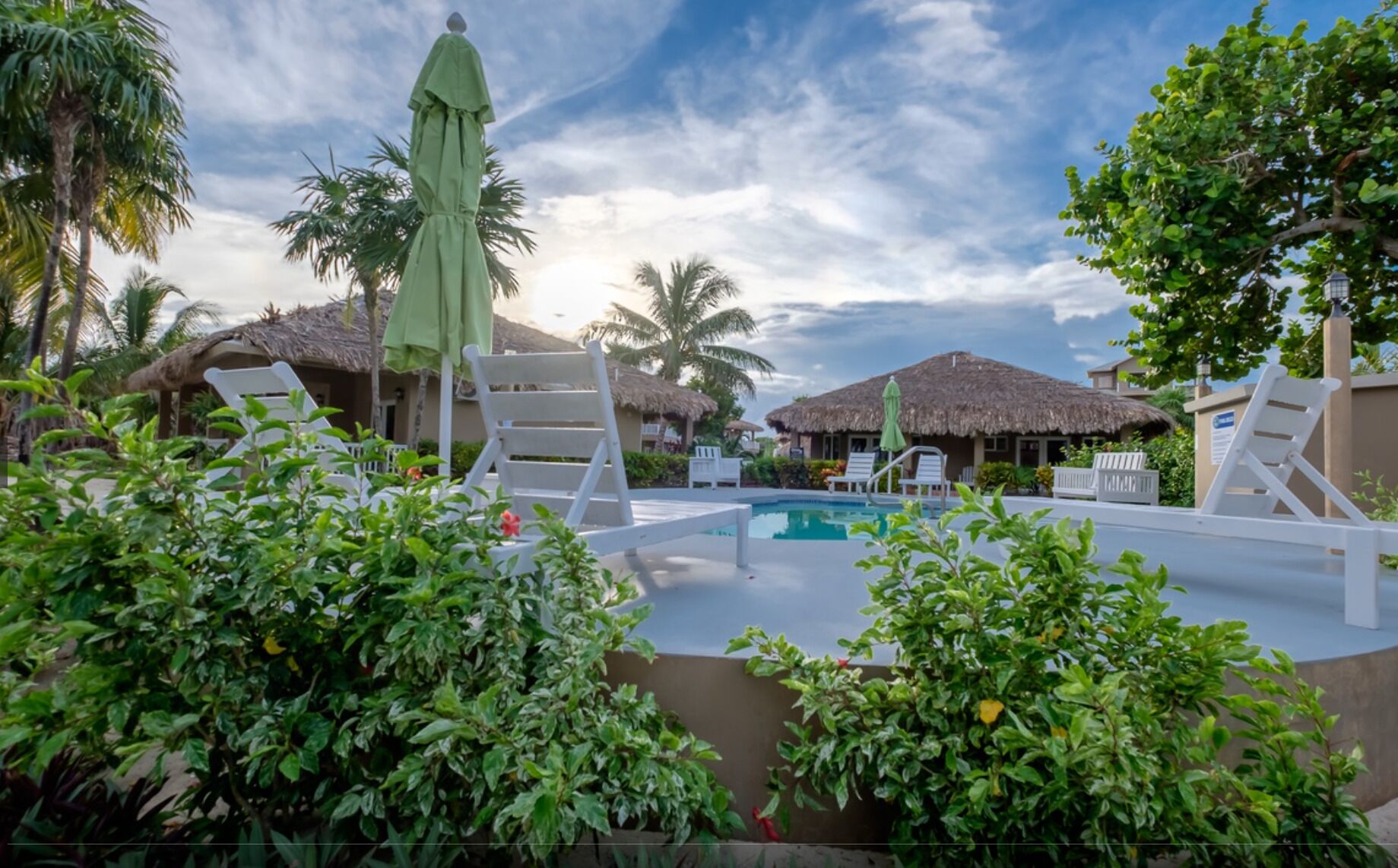 A pool area with lounge chairs, a thatched-roof structure, and lush greenery.