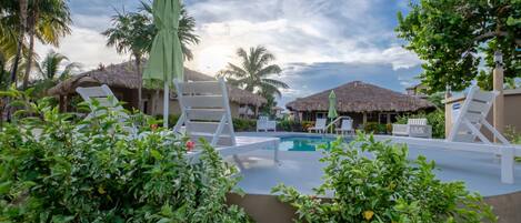 A pool area with lounge chairs, a thatched-roof structure, and lush greenery.