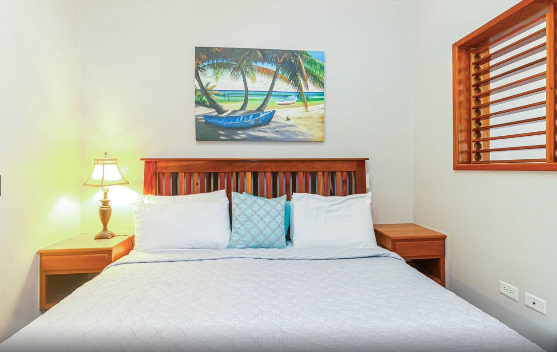 A neatly made bed with a wooden headboard, a bedside lamp, and a painting of a beach scene.