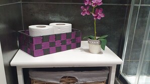 Ein Badezimmer-Waschtisch mit einer weißen Ablagefläche, auf der sich ein violett-schwarzer Schachbrettkorb, eine Toilettenpapierrolle und eine Topforchidee befinden.