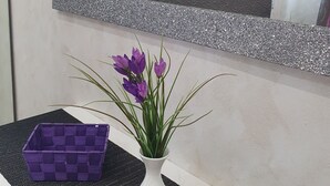 Ein Badezimmer mit Spiegel, einer Vase mit violetten Blumen und einem violetten Korb auf einer strukturierten Arbeitsfläche.