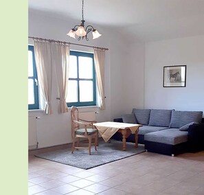 A living room with a sofa, a table, and a chair. There are two windows with curtains, a chandelier, and a framed picture on the wall. - Apartment 7 equestrian facility - riding and holiday farm Blohm - object 26099 (Rostock)