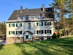 Un bâtiment à deux étages, doté de fondations en pierre, de nombreuses fenêtres avec volets et d’un petit porche avec un banc.