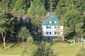 A two-story house with a green roof and white walls, surrounded by dense trees and a grassy field.