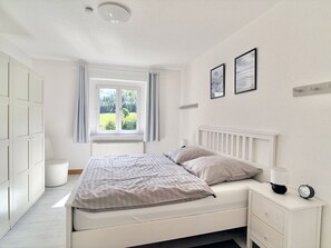 A bedroom with a bed, bedside table, lamp, clock, and a window with curtains.