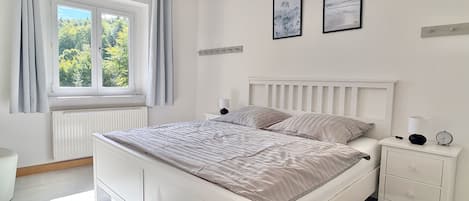 A bedroom with a bed, a bedside table with a lamp, a clock, and two framed pictures on the wall.