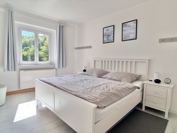 A bedroom with a bed, a bedside table with a lamp, a clock, and two framed pictures on the wall.