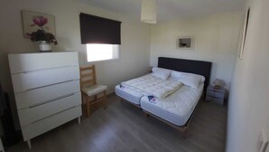 A bedroom with a bed, a dresser, a chair, a window with a blind, a bedside table, and a wall-mounted light fixture. - In the city or in the countryside! 2.7 km from the Valentré Bridge (PRADINES)