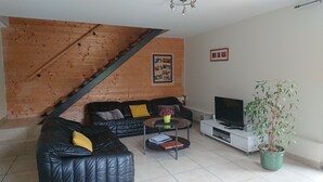 A living room with a black leather sofa, a coffee table with a book and a vase, a TV on a white stand, and a staircase against the wall. - In the city or in the countryside! 2.7 km from the Valentré Bridge (PRADINES)