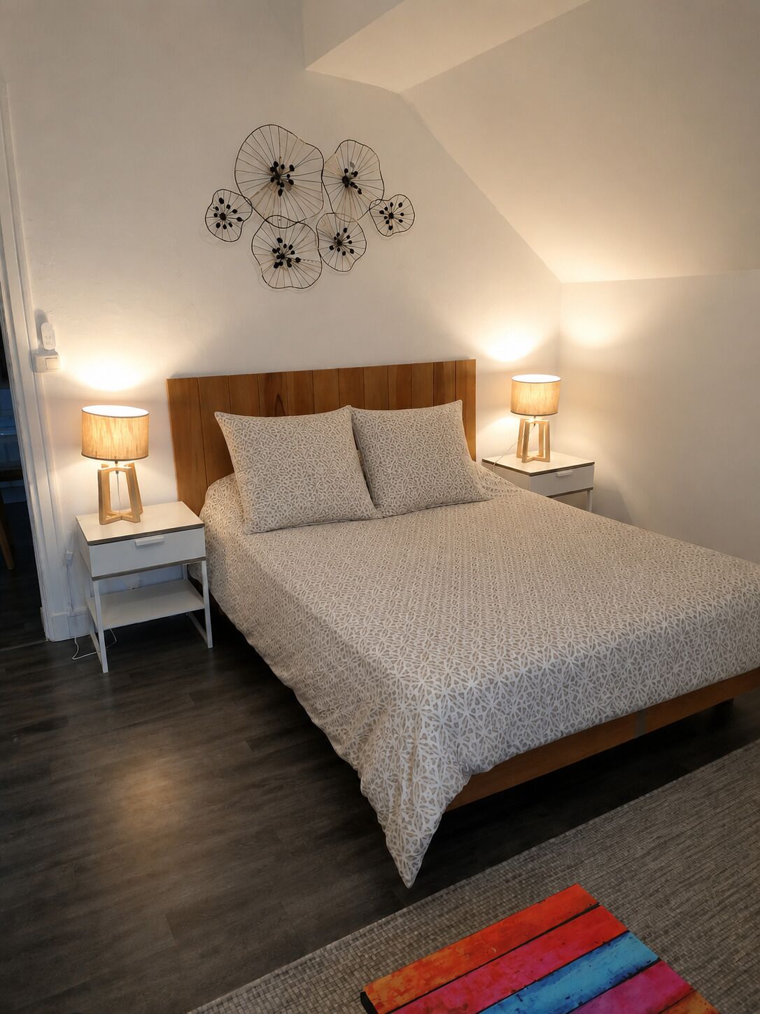 Een slaapkamer met een houten hoofdbord, twee nachtkastjes met lampen, een bed met een geborduurde spreideken en een bloemendecoratie aan de muur.