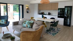 Un salon moderne avec un canapé, des fauteuils et un coin repas. La cuisine est équipée de meubles blancs et d’un réfrigérateur noir.