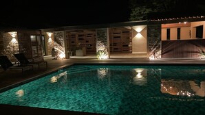 Une piscine nocturne à l’eau claire, entourée d’une terrasse en bois et de murs en pierre.