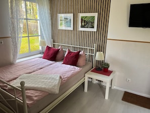 A bedroom with a bed, pillows, a bedside table with a lamp, a TV, and a window with curtains.