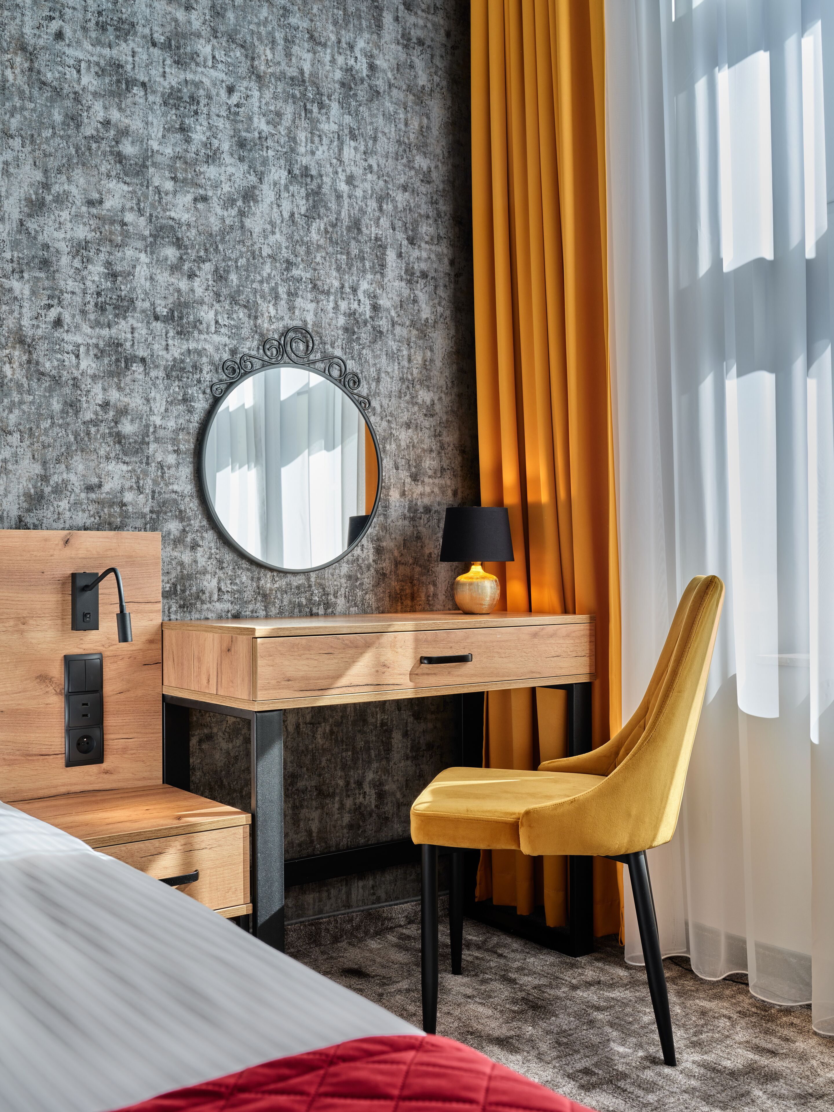 Een moderne hotelkamer met een houten bureau, een ronde spiegel, een lamp, een gele stoel en een bed met rode accenten.