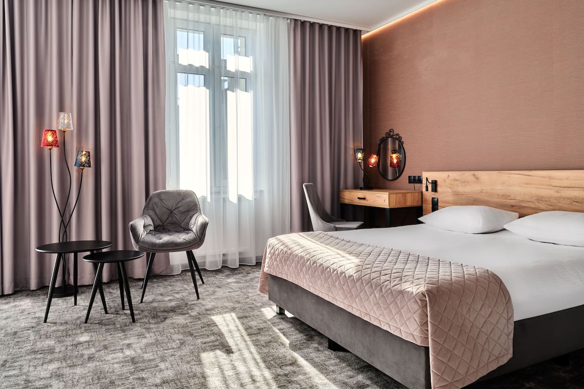 A hotel room with a bed, two chairs, a desk, a mirror, and a window with curtains.