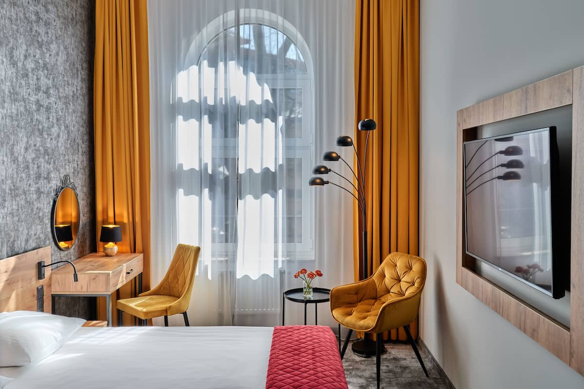 A hotel room with a bed, a desk with a lamp, a chair, a TV, and a window with curtains.