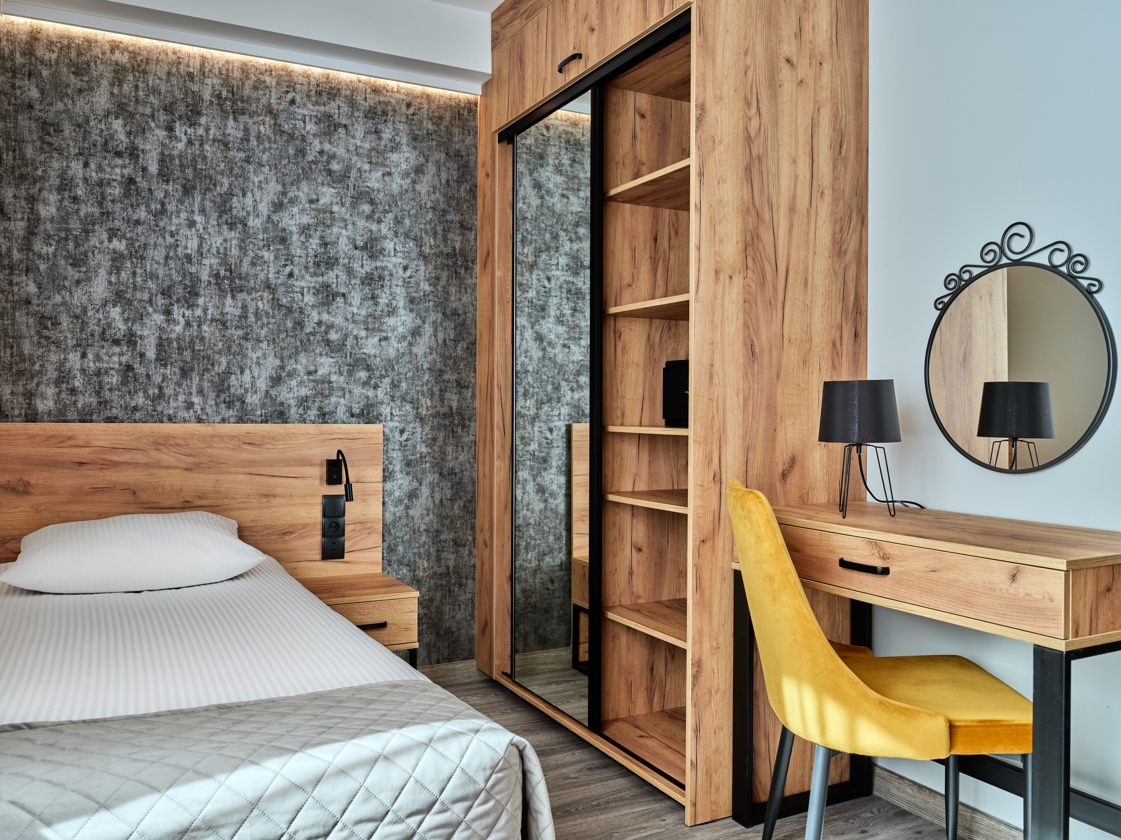 A bedroom with a bed, a wooden wardrobe, a desk with a chair, and a mirror.