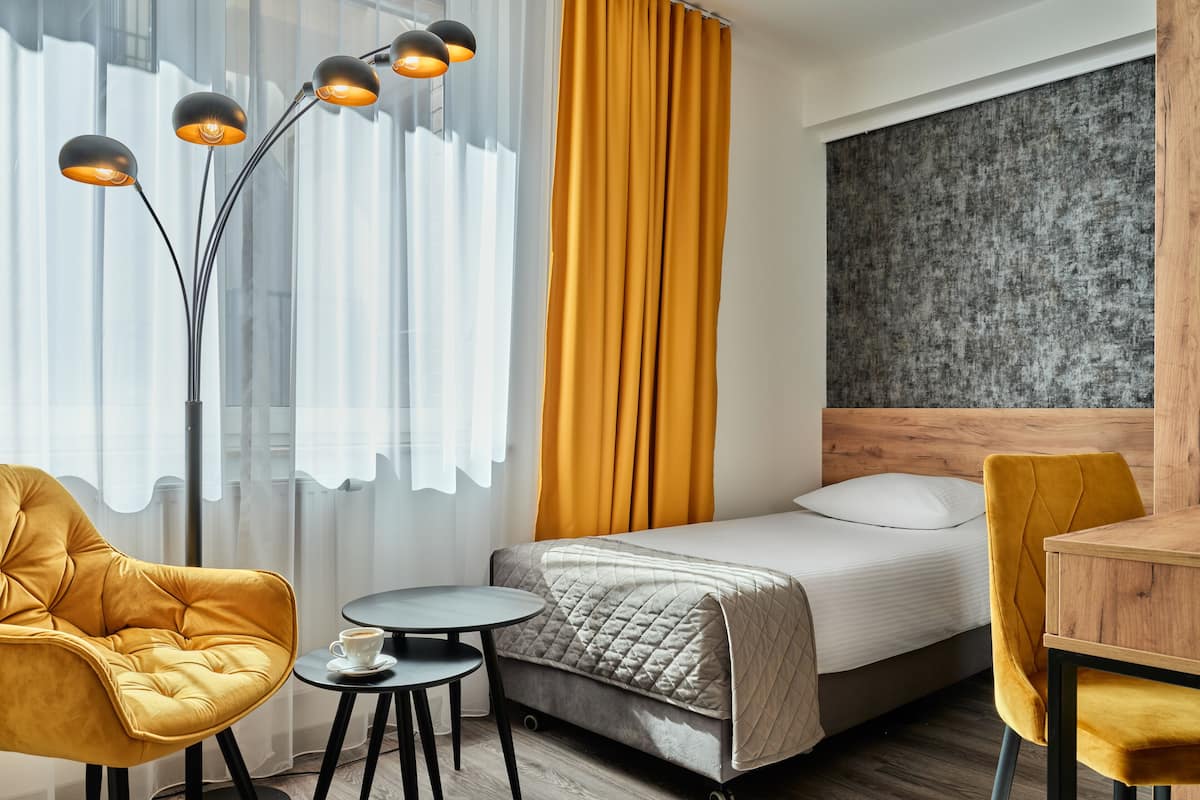 A modern hotel room with a bed, a round table, a chair, a floor lamp, and a view of the outside.