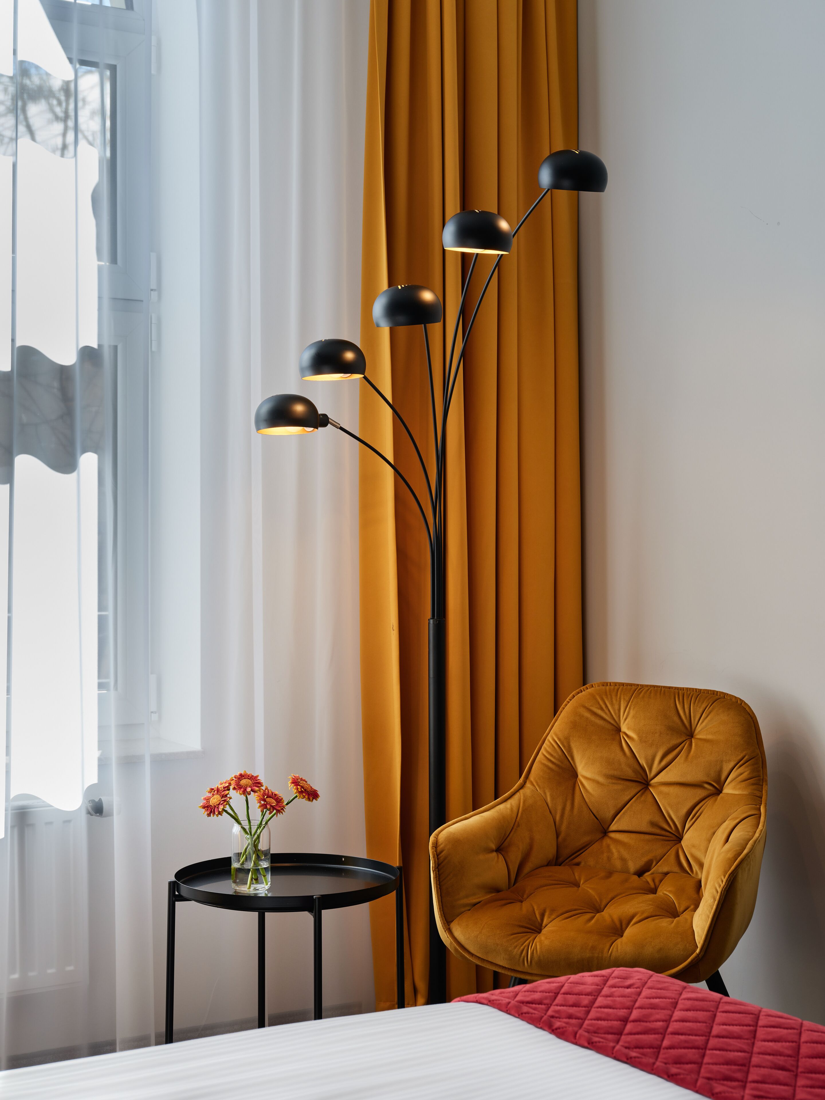 A room with a bed, a yellow armchair, a floor lamp, and a small table with flowers.