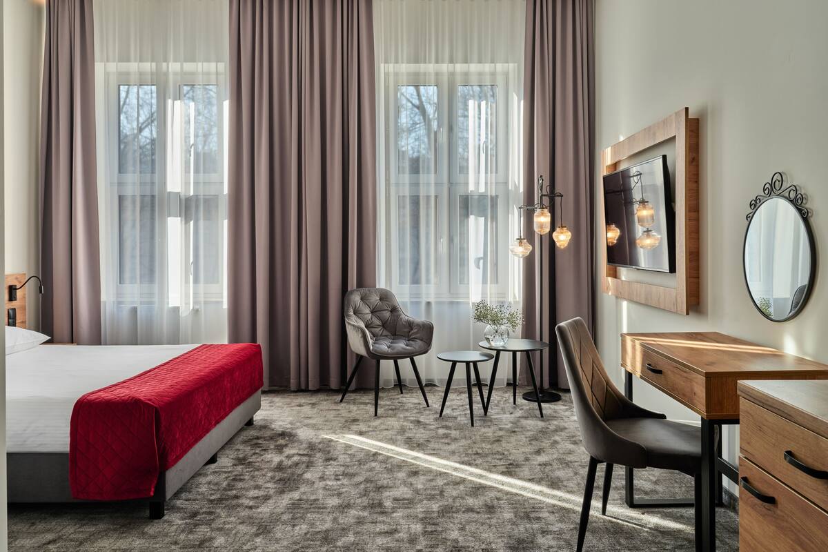 A hotel room with a bed, a desk, a chair, a small table, and a mirror.