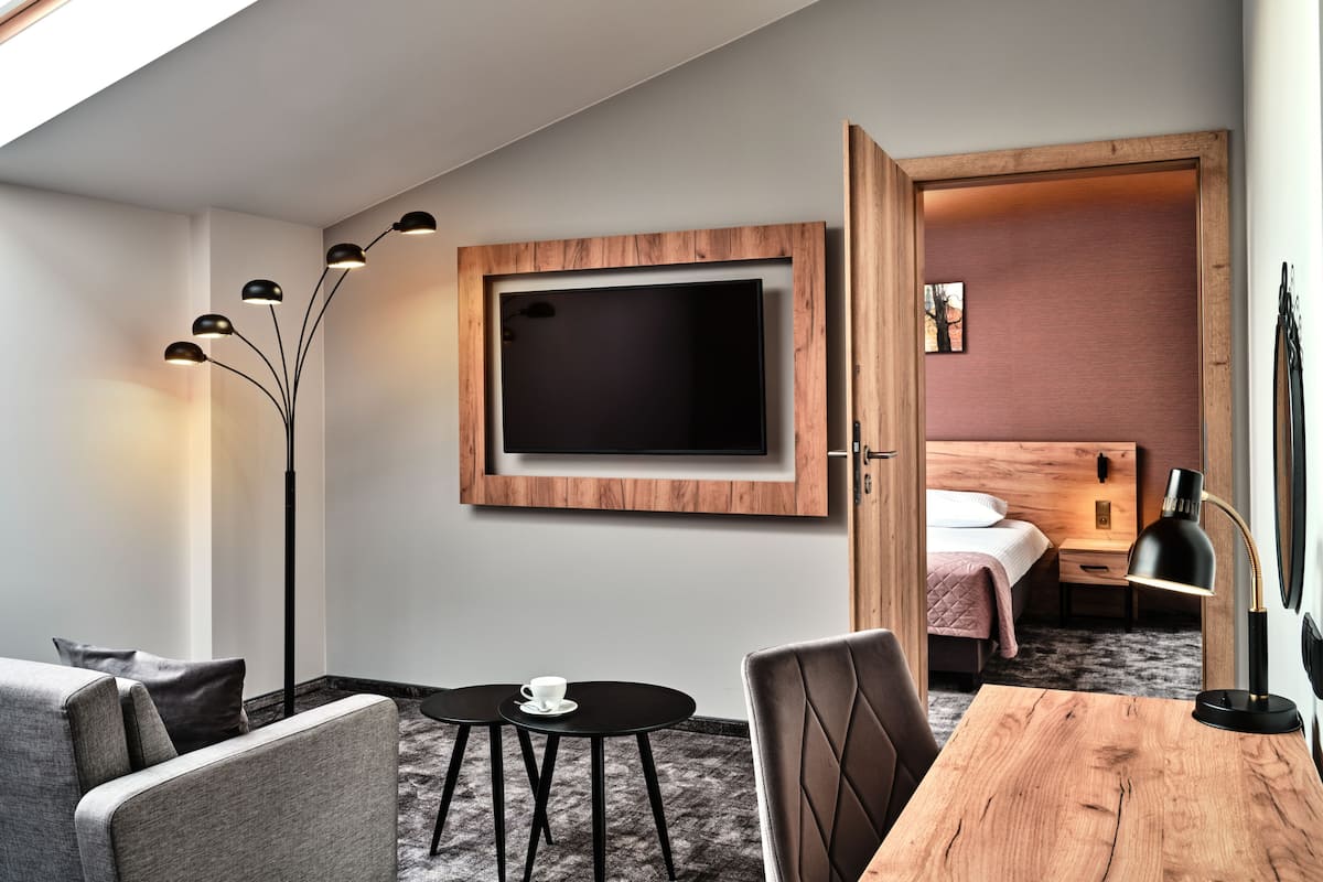 A modern hotel room with a flat-screen TV mounted on a wooden wall, a dining table with chairs, a small round table with a cup, a sofa, and a bed with a wooden headboard.