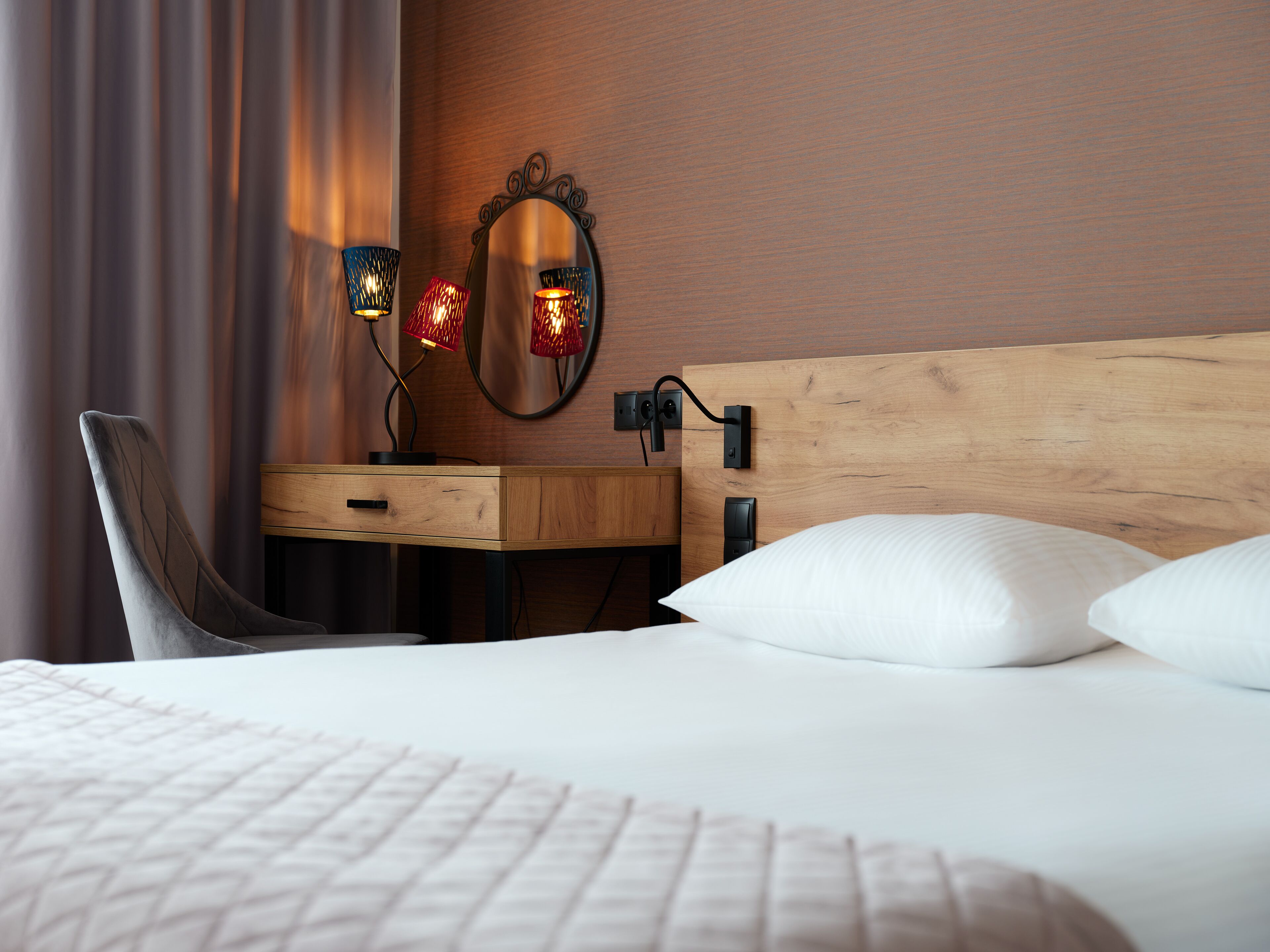 A neatly arranged hotel room with a wooden headboard, a bedside table with a lamp, a chair, and a large mirror.
