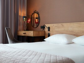 Una habitación de hotel ordenada con cabecera de madera, una mesita de noche con lámpara, una silla y un espejo grande.