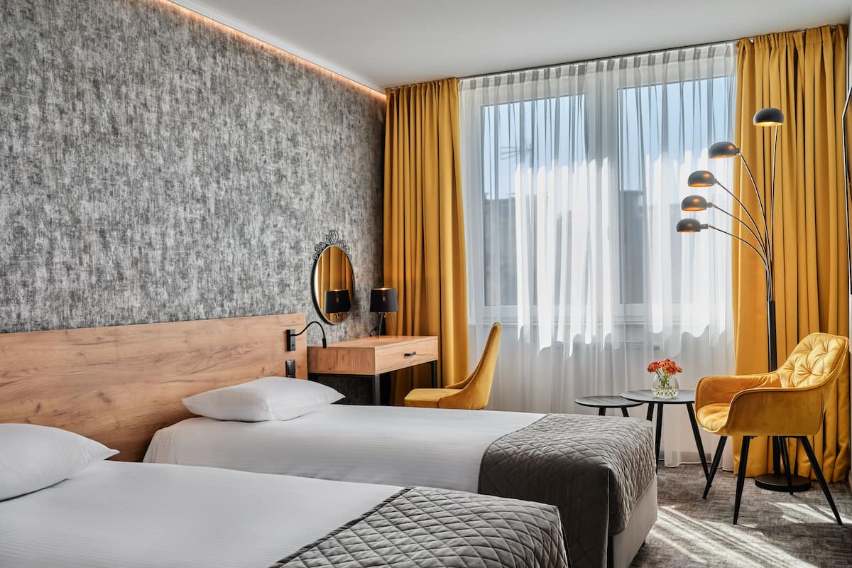 A hotel room with two beds, a wooden headboard, a desk with a mirror, a chair, and a small table with a vase.