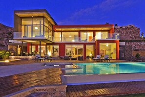 A modern two-story house with a swimming pool, outdoor seating, and a stone wall backdrop.