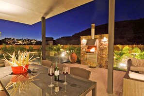 An outdoor patio with a stone fireplace, wicker furniture, and a glass table with wine glasses and a bottle.