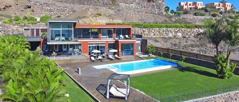 A modern two-story house with a swimming pool, a wooden deck, and a view of a hillside with other houses.