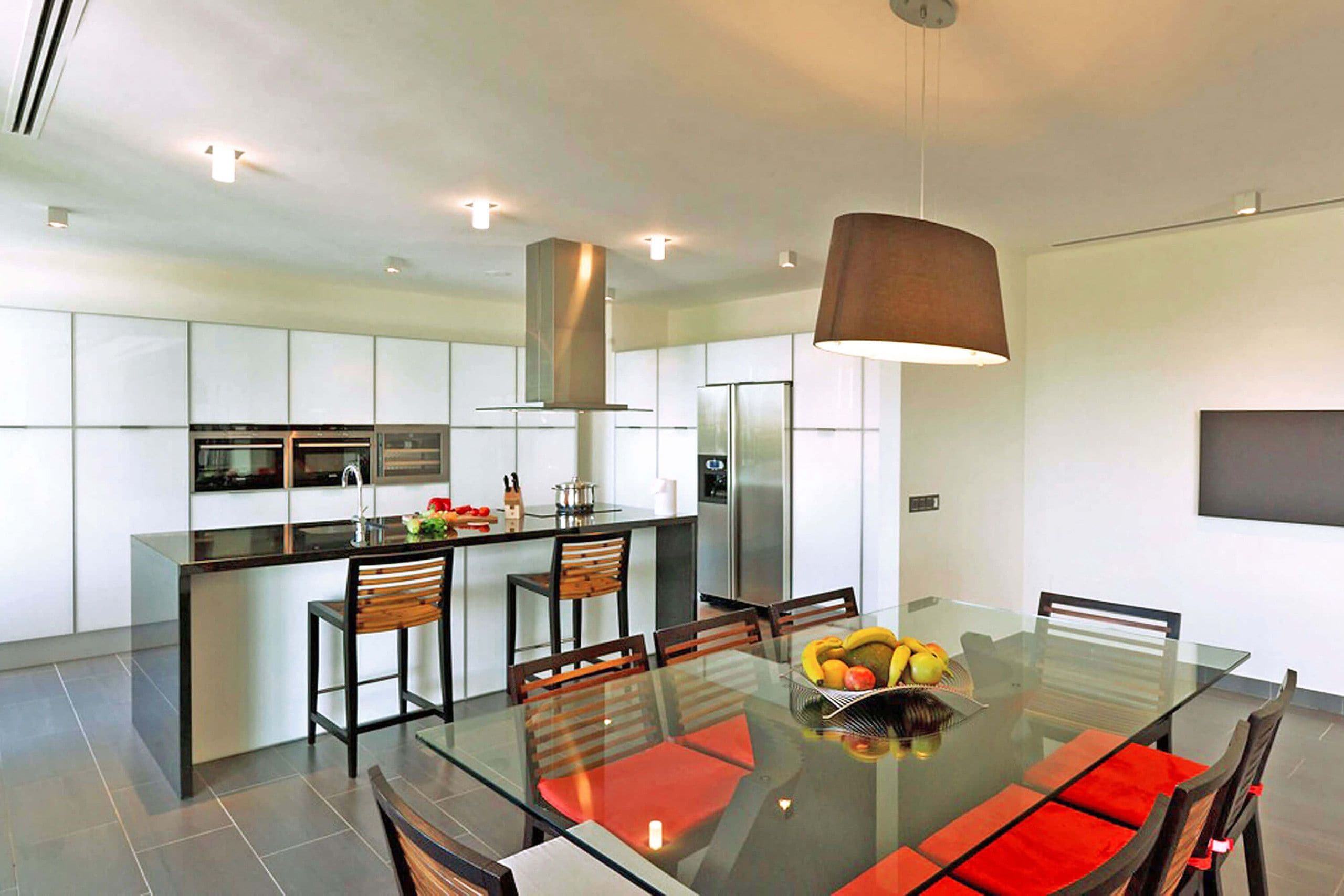 A modern kitchen with a central island, stainless steel appliances, and a dining area with a glass table and red cushions.