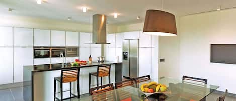 A modern kitchen with a central island, stainless steel appliances, and a dining area with a glass table and red cushions.
