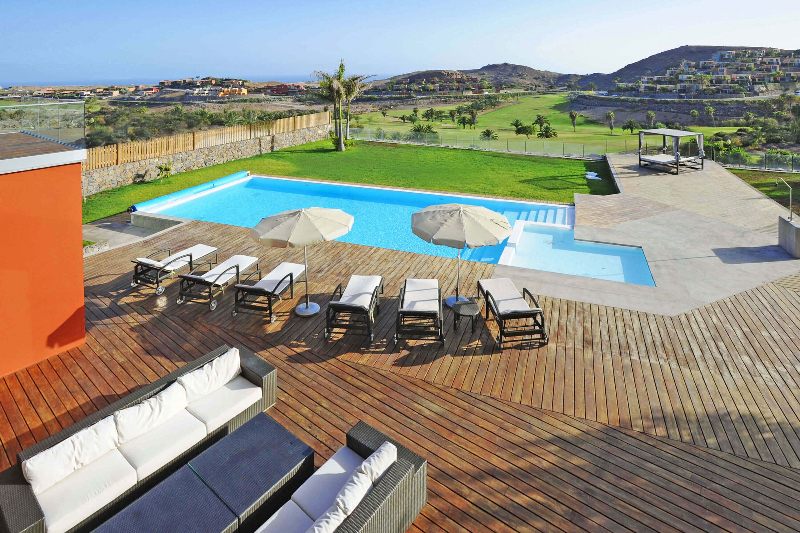 Zona de piscina con sillones de descanso y sombrillas, una terraza de madera y vistas a un campo de golf y colinas al fondo.