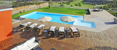 Zona de piscina con sillones de descanso y sombrillas, una terraza de madera y vistas a un campo de golf y colinas al fondo.