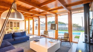 Una sala de estar moderna con un sofá, una mesa de centro y sillas, grandes ventanales con vista a una piscina y zona exterior.