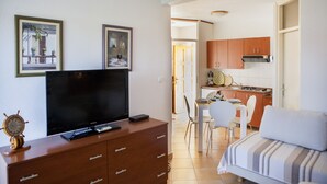 A living room with a TV, wooden cabinet, and a sofa. There is a dining area with chairs and a table.