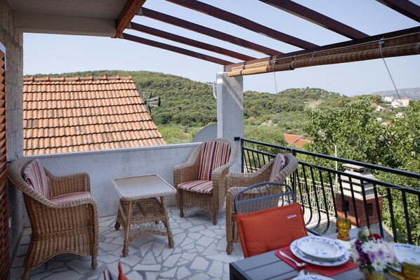 A balcony with wicker furniture, a table set for dining, and a view of a hilly landscape.
