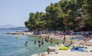 A crowded beach with people swimming and sunbathing, surrounded by trees and rocky terrain.