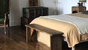 A bedroom with a wooden bed, a bench, a green chair, a wooden nightstand, and a wall-mounted lamp.