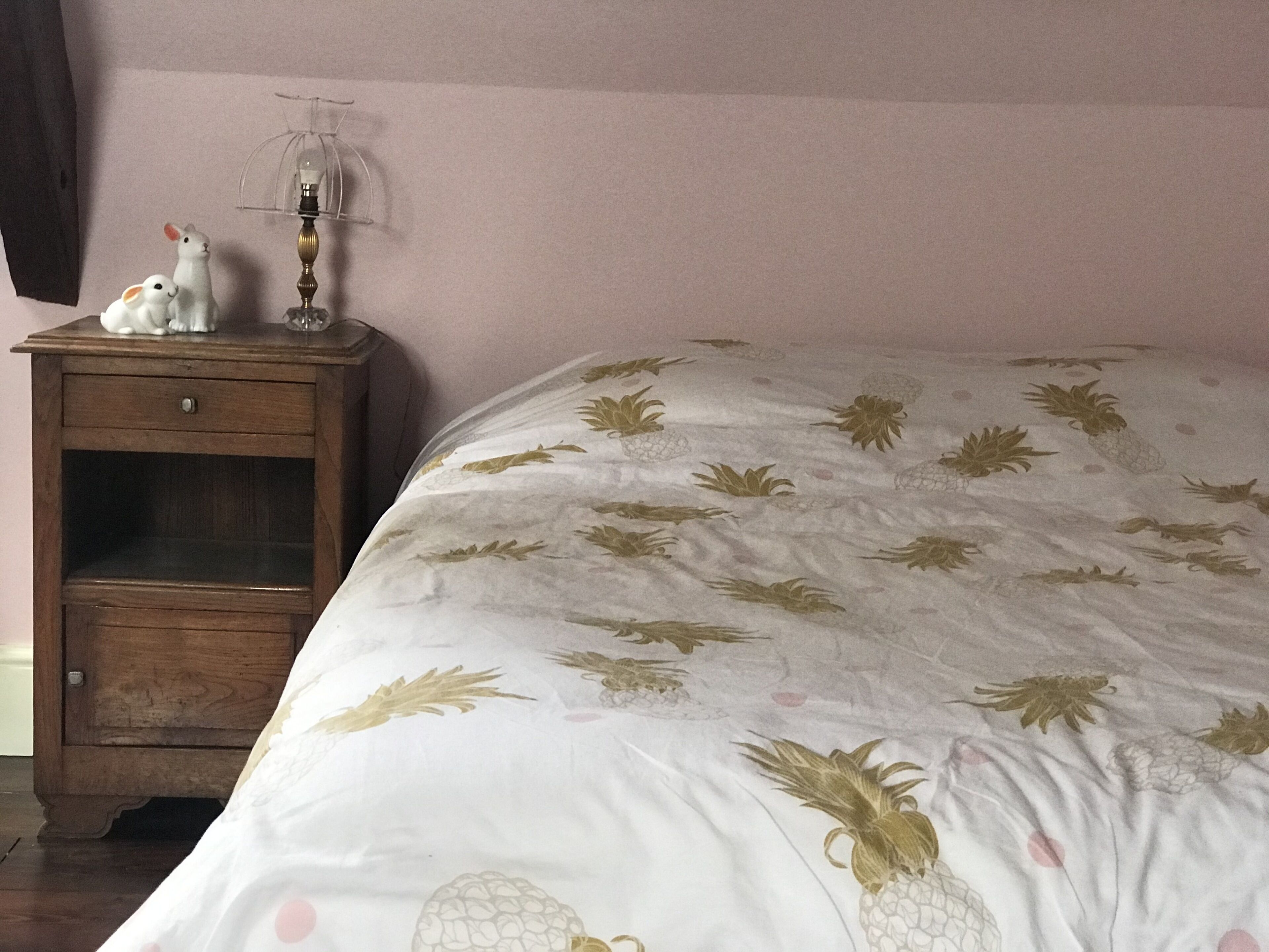 A bedroom with a bed featuring a pineapple pattern, a wooden nightstand with a lamp, and two white figurines.