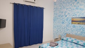 A bedroom with a bed, a blue curtain, a wall-mounted air conditioner, a nightstand, a remote control, and a painting on the wall.