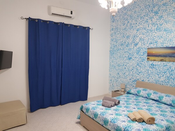 A bedroom with a bed, a blue curtain, a wall-mounted air conditioner, a nightstand, a remote control, and a painting on the wall.