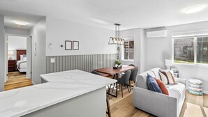 Una sala de estar moderna con un sofá gris, una mesa de comedor de madera y una zona de cocina con encimera blanca.