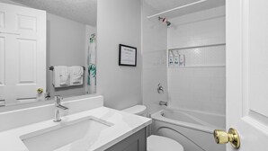 Un baño moderno con lavamanos, inodoro y bañera blancos. Hay un cartel enmarcado en la pared y un perchero con toallas.