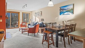 A spacious living room with a large window, a dining area with wooden furniture, and a painting on the wall. - C303 Peaceful Canmore Corner- Downtown, Free parking, View, 2BR, 2BT, Sleeps 7 (Canmore)